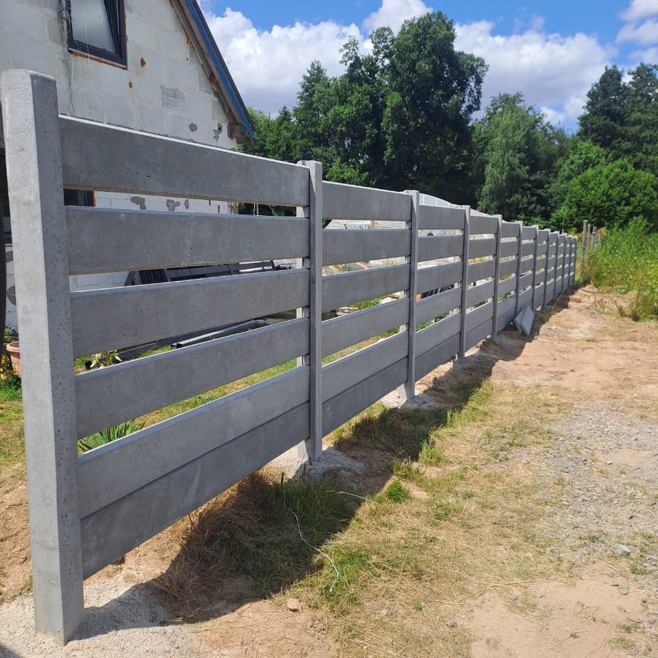 Nowoczesne ogrodzenie betonowe z poziomymi panelami w odcieniach szarości, na tle domu w budowie i zieleni. Widok z perspektywy ziemi, słoneczny dzień.