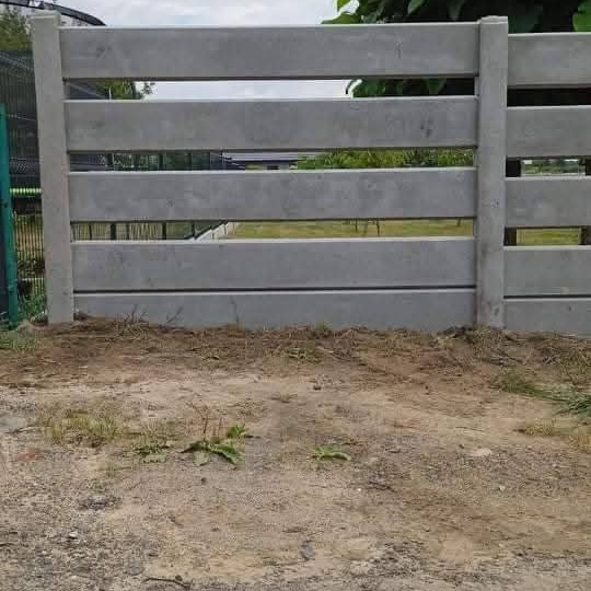 Szary, prefabrykowany płot betonowy z poziomymi przęsłami, widoczny z bliska na tle ziemi i zieleni. Element ogrodzenia, montaż.