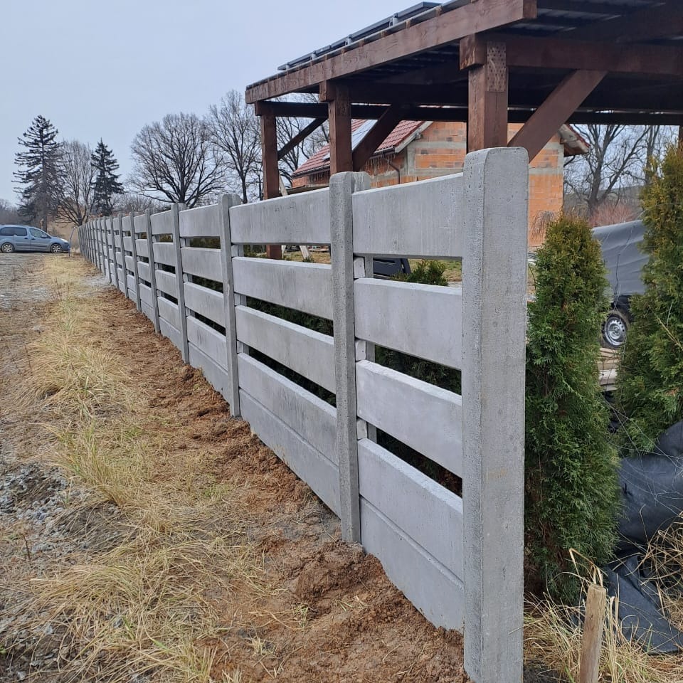 Nowoczesne ogrodzenie betonowe z poziomymi szczelinami, świeżo zamontowane, z widoczną ziemią i zielonymi tujami w tle, pod pochmurnym niebem.