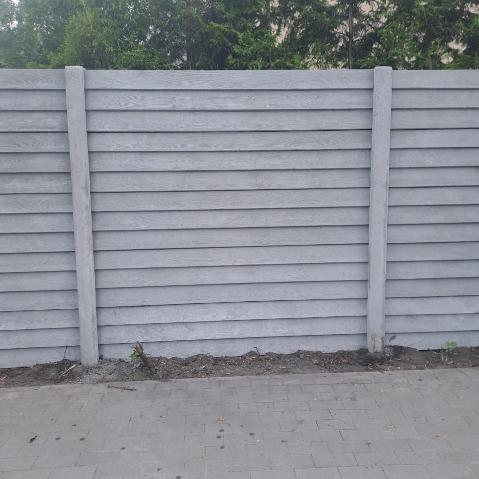 Szary, poziomy panel ogrodzeniowy z betonowymi słupkami, imitujący drewno, z zielonymi drzewami w tle i szarą kostką brukową u dołu.