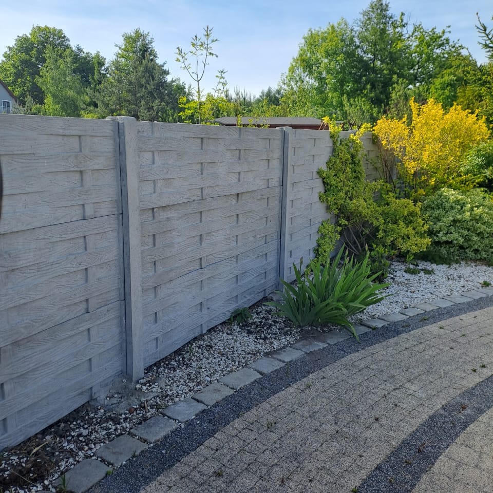 Szary, betonowy płot o teksturze plecionki na tle zieleni ogrodu. Widoczny fragment ścieżki z kostki brukowej i kamyczków. Słoneczny dzień.