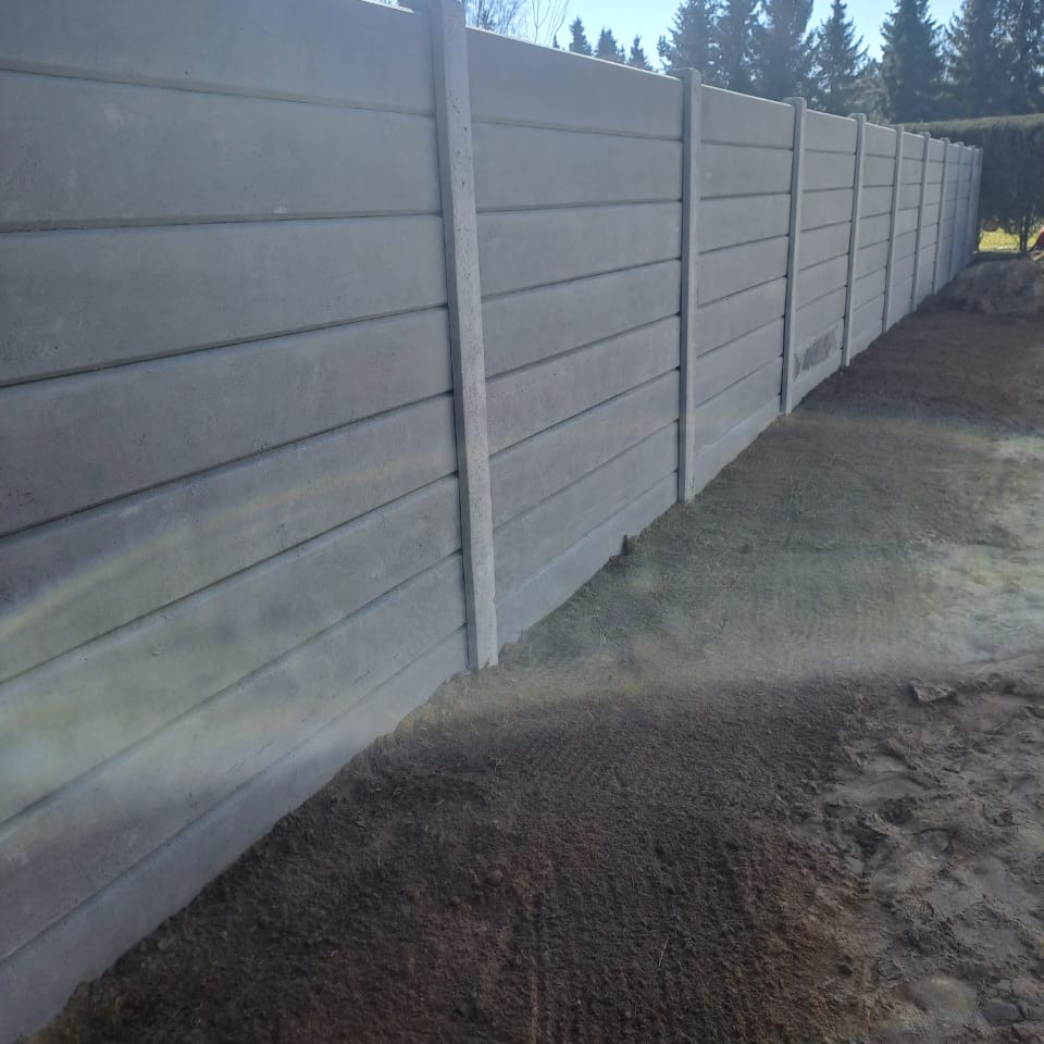 Długi, szary, betonowy płot panelowy z poziomymi deskami, widziany pod kątem, z ziemią u podstawy i drzewami w tle. Minimalistyczny styl.