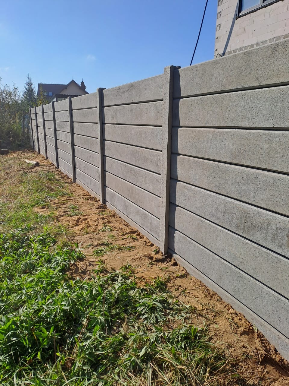 Szary, prefabrykowany płot betonowy z poziomymi panelami, oddzielający posesję. Widoczne słupki i panele, trawa i piasek u podstawy, dom w tle.