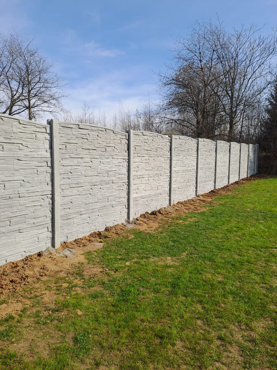 Długi, szary, betonowy płot o fakturze kamienia oddziela trawnik od drzew. Widoczne słupki i panele. Płot jest nowy, a trawa zielona. Słoneczny dzień.