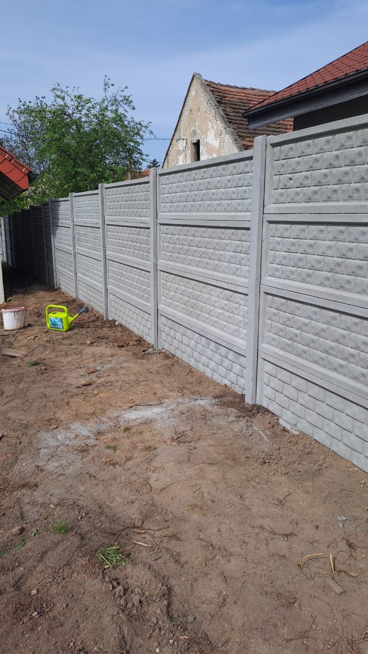 Szary, betonowy płot z wzorem cegły oddziela posesję. Obok płotu leży zielona konewka i białe wiadro. W tle dom z czerwonym dachem i drzewo.