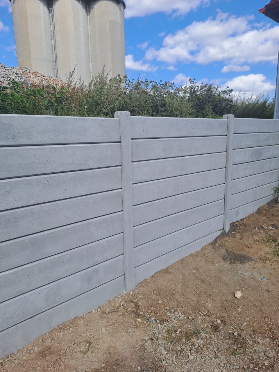 Nowoczesne ogrodzenie betonowe z poziomych paneli, widoczne na tle błękitnego nieba i przemysłowego krajobrazu z silosami w oddali.