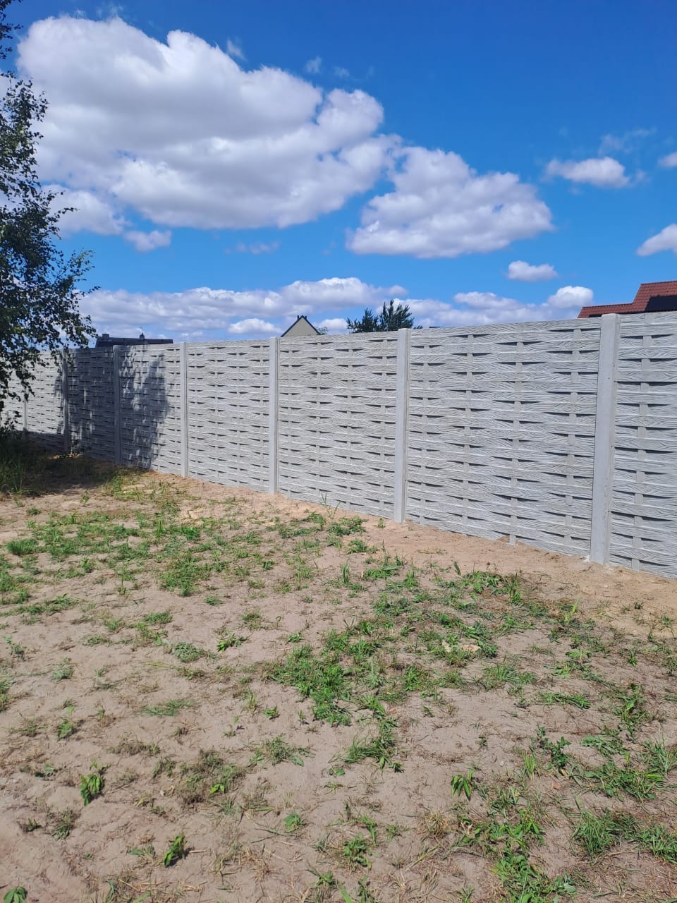 Nowoczesne, ażurowe ogrodzenie betonowe w kolorze szarym, oddzielające posesję. Widok na błękitne niebo z białymi chmurami w słoneczny dzień.