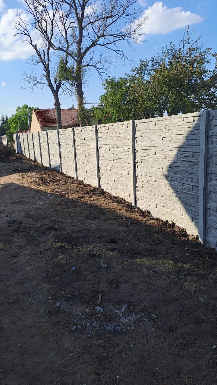 Długi, szary ogrodzenie betonowe z fakturą kamienia, rozciągające się wzdłuż nierównego terenu z widocznymi drzewami i fragmentem budynku w tle.