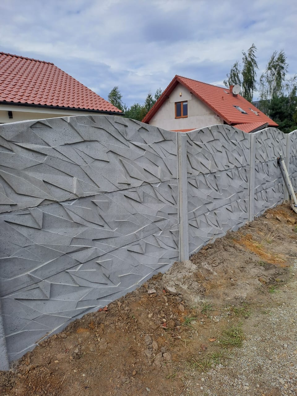 Szary, betonowy płot o geometrycznym wzorze, oddzielający posesję. W tle widoczne dwa domy z czerwoną dachówką i błękitne niebo.