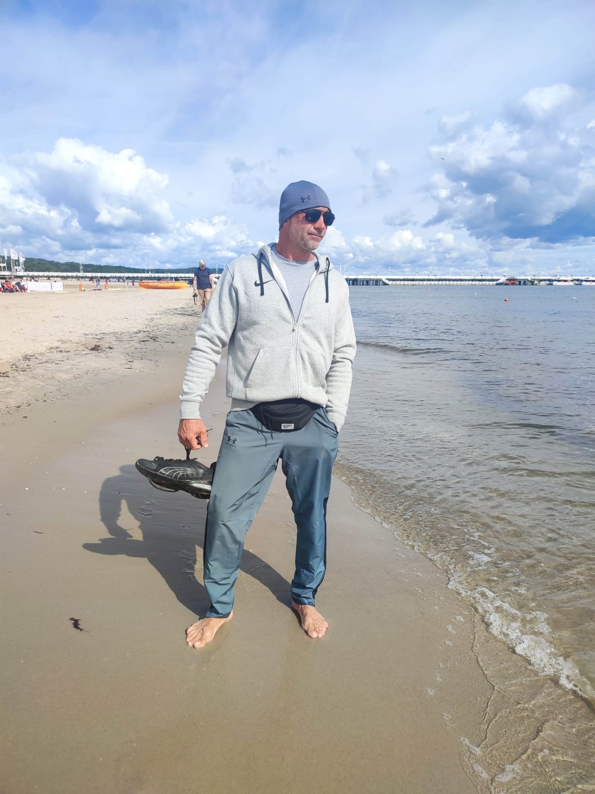 Mężczyzna na plaży w Lubockiem, trzymający buty w ręku, w tle morze i molo, pochmurne niebo. Casualowy strój, relaks w nadmorskim krajobrazie.