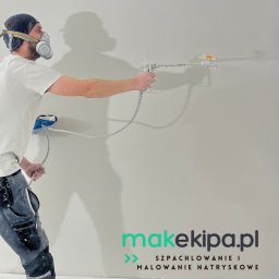 MAK - KONSERWACJA ZABYTKÓW, PRACE OGÓLNOBUDOWLANE SPÓŁKA Z OGRANICZONĄ ODPOWIEDZIALNOŚCIĄ - Pracownik w masce maluje ścianę natryskowo, używając pistoletu. Widoczny fragment ściany, przewód i dolna część ciała malarza w ubrudzonych farbą spodniach.