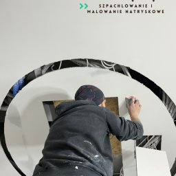 MAK - KONSERWACJA ZABYTKÓW, PRACE OGÓLNOBUDOWLANE SPÓŁKA Z OGRANICZONĄ ODPOWIEDZIALNOŚCIĄ - Pracownik szlifuje gładź na suficie z nietypowym okrągłym otworem, widoczne kable i instalacje. Szpachelka w kieszeni, logo firmy w tle. Praca w trakcie wykończenia.