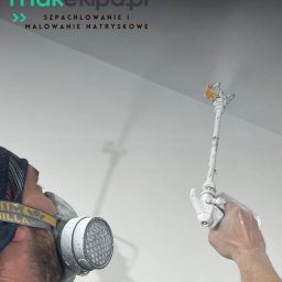 MAK - KONSERWACJA ZABYTKÓW, PRACE OGÓLNOBUDOWLANE SPÓŁKA Z OGRANICZONĄ ODPOWIEDZIALNOŚCIĄ - Malowanie natryskowe sufitu przez pracownika w masce ochronnej. Widoczny pistolet natryskowy i świeżo pomalowana, gładka powierzchnia. Praca w toku.