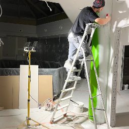 MAK - KONSERWACJA ZABYTKÓW, PRACE OGÓLNOBUDOWLANE SPÓŁKA Z OGRANICZONĄ ODPOWIEDZIALNOŚCIĄ - Pracownik na drabinie szpachluje ścianę w jasnym wnętrzu. Widoczna lampa budowlana, narzędzia i sufit z instalacją. Etap prac wykończeniowych w pomieszczeniu.