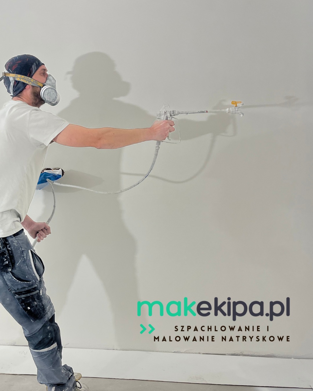 Pracownik w masce maluje ścianę natryskowo, używając pistoletu. Widoczny fragment ściany, przewód i dolna część ciała malarza w ubrudzonych farbą spodniach.