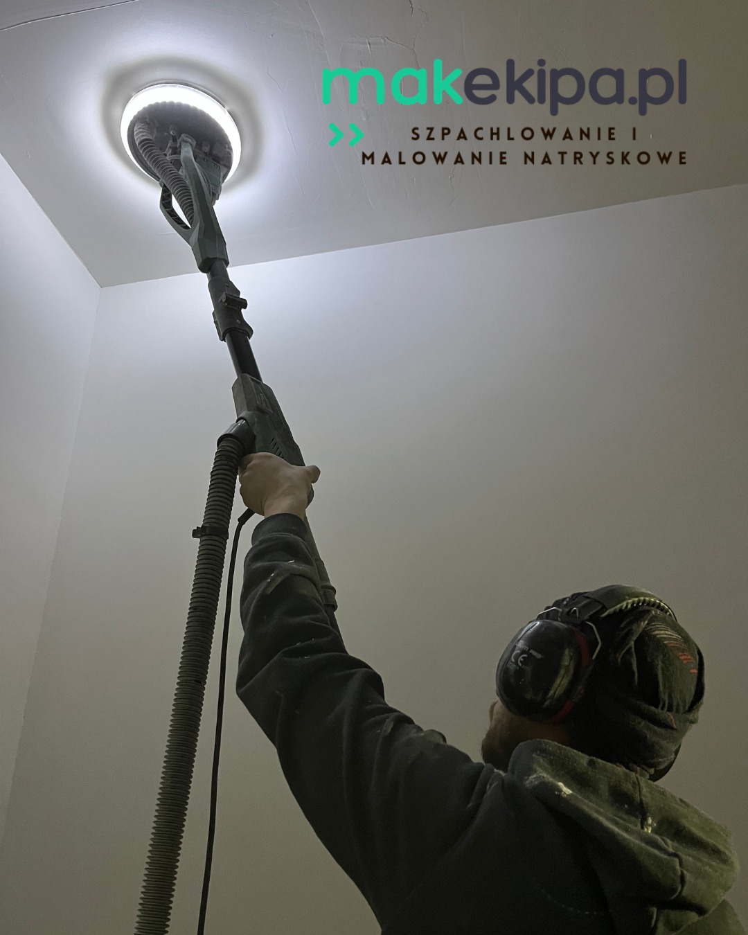 Pracownik w słuchawkach szlifuje sufit szlifierką z oświetleniem, widoczne logo firmy i informacja o szpachlowaniu i malowaniu natryskowym. Ujęcie od dołu.