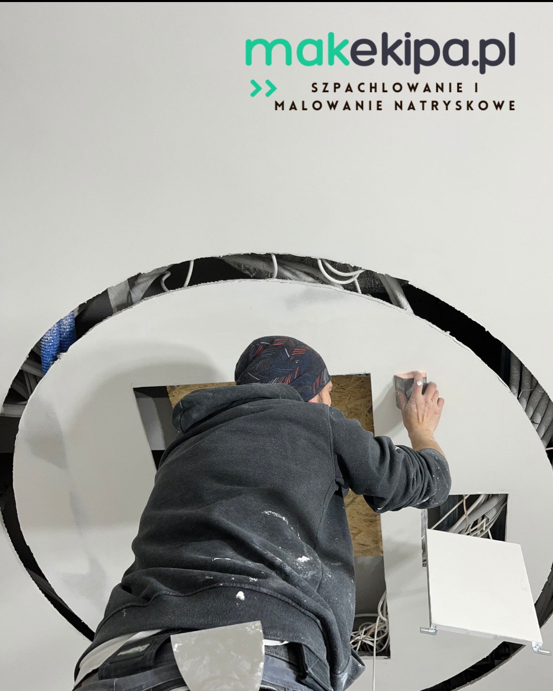 Pracownik szlifuje gładź na suficie z nietypowym okrągłym otworem, widoczne kable i instalacje. Szpachelka w kieszeni, logo firmy w tle. Praca w trakcie wykończenia.