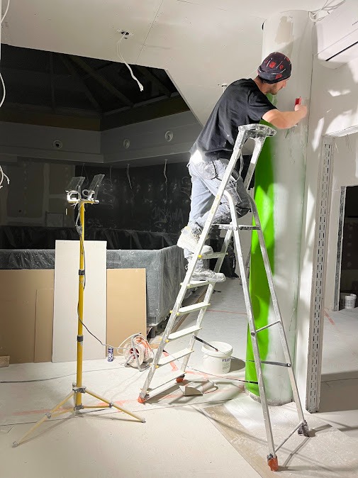 Pracownik na drabinie szpachluje ścianę w jasnym wnętrzu. Widoczna lampa budowlana, narzędzia i sufit z instalacją. Etap prac wykończeniowych w pomieszczeniu.