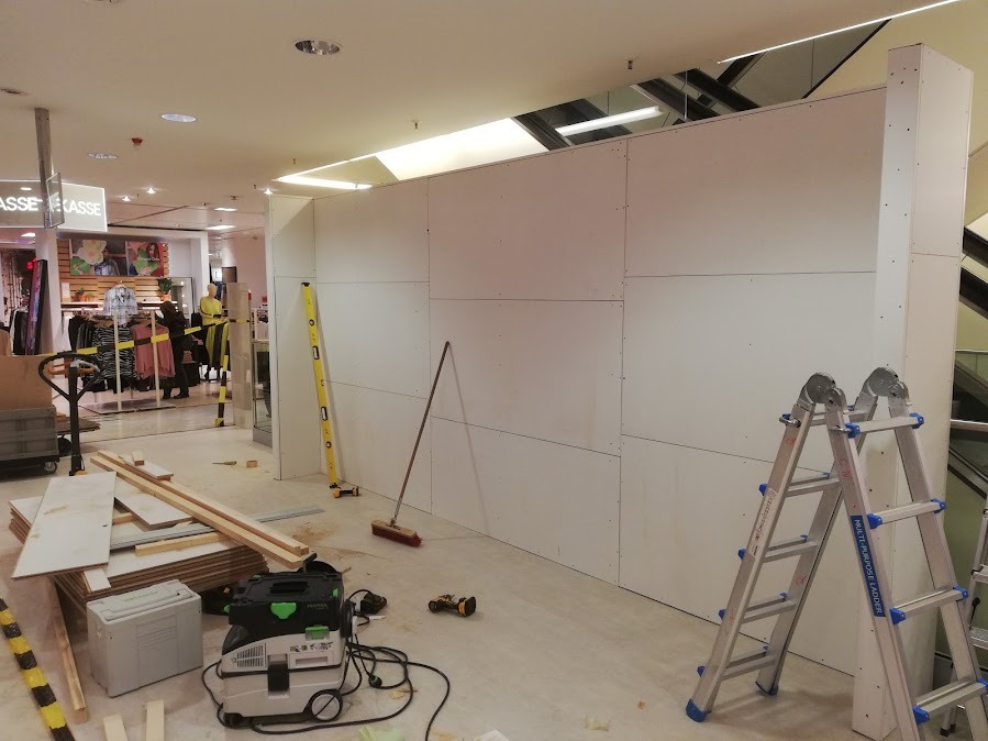 Montaż ścianki działowej z płyt gipsowo-kartonowych w przestrzeni handlowej. Widoczne narzędzia, drabina i poziomica. Prace w toku, surowy stan wykończenia.