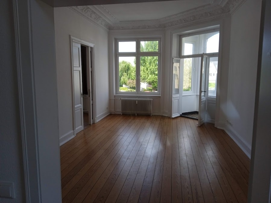 Jasne, przestronne wnętrze po remoncie: parkiet, białe ściany z dekoracyjną sztukaterią, okna z widokiem na zieleń. Drzwi wewnętrzne i balkonowe otwarte, wpuszczające światło.