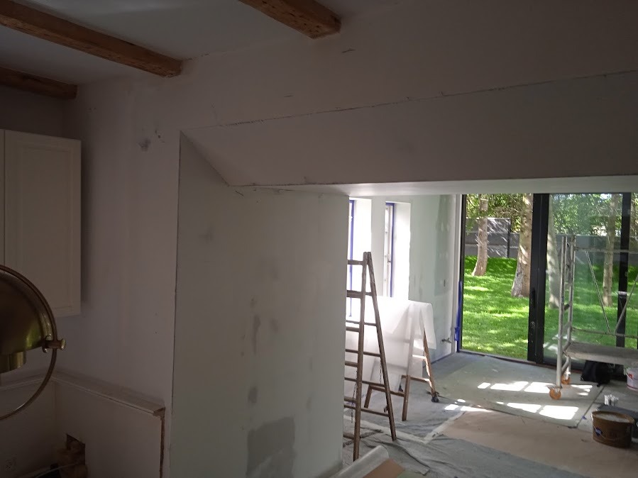 Wnętrze w trakcie prac wykończeniowych: ściany przygotowane do malowania, widoczne ślady szpachlowania, drabina i zabezpieczona podłoga. W tle widok na ogród.