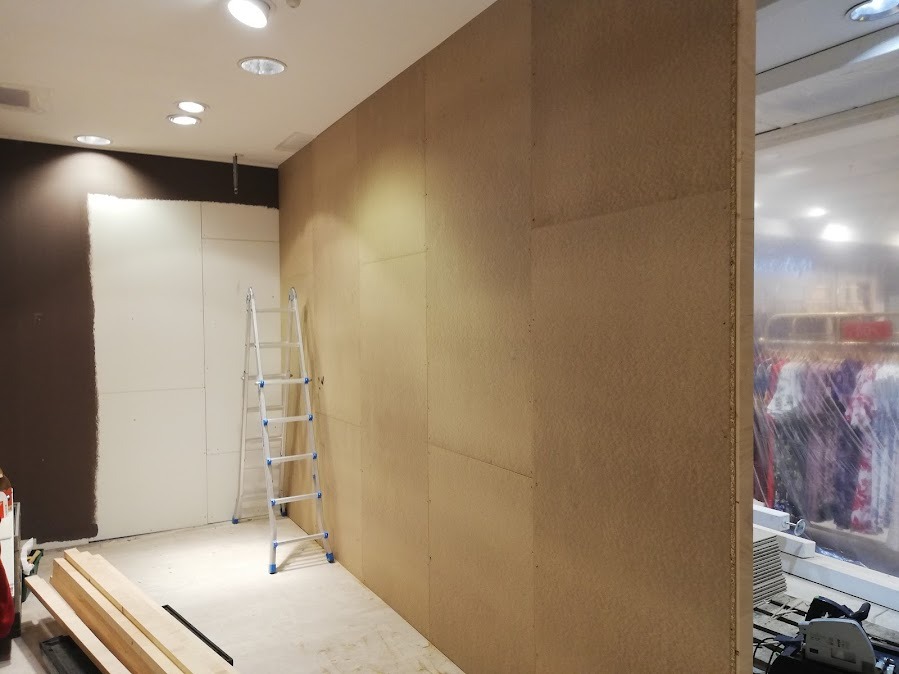 Montaż płyt na ścianie w lokalu usługowym. Widoczna drabina, płyty w kolorze jasnego drewna, częściowo pomalowana ściana. Prace wykończeniowe w toku.