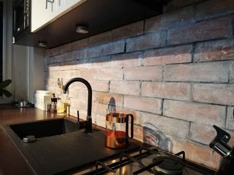 Kuchnia z ceglaną ścianą nad blatem i czarnym zlewem. Widoczne oświetlenie podszafkowe i akcesoria kuchenne. Cegła w odcieniach bieli i czerwieni tworzy rustykalny klimat.
