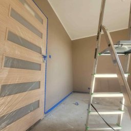 Mateos - Pokój w trakcie malowania na beżowo, zabezpieczone listwy podłogowe taśmą, drzwi oklejone folią, widoczna drabina malarska.