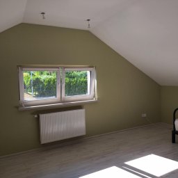 Mateos - Poddasze ze ścianą w odcieniach zieleni, grzejnikiem pod oknem z roletami wewnętrznymi i jasną podłogą. Widoczne promienie słoneczne na podłodze.