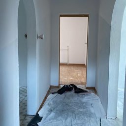 Mateos - Korytarz w trakcie malowania: świeżo pomalowane ściany na biało, podłoga zabezpieczona folią malarską, widoczne łuki w przejściach i otwarte drzwi do pokoju.