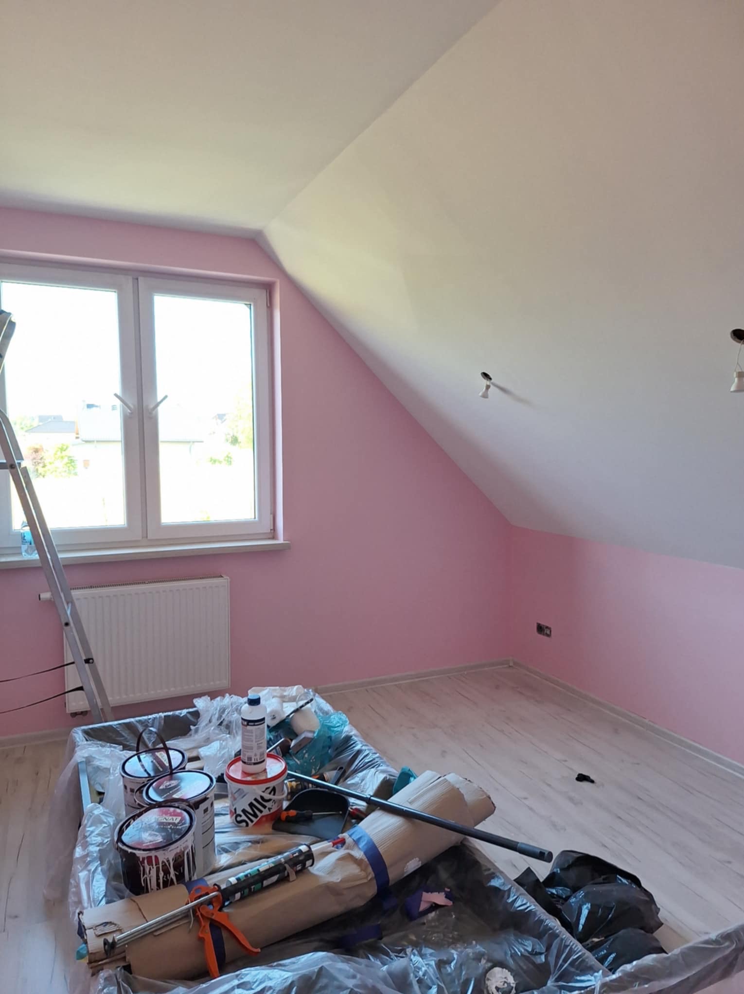 Pokój na poddaszu po malowaniu ścian na delikatny róż, z widocznymi narzędziami i farbami, drabina oparta o okno, jasna podłoga.