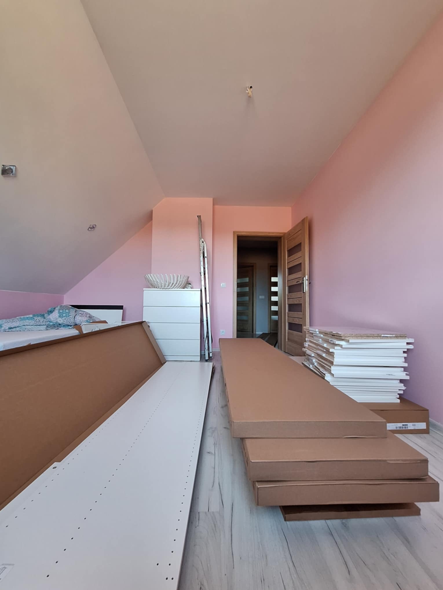 Pokój na poddaszu z przygotowanymi elementami do montażu mebli. Widoczne kartony i białe płyty. W tle komoda i drabina.