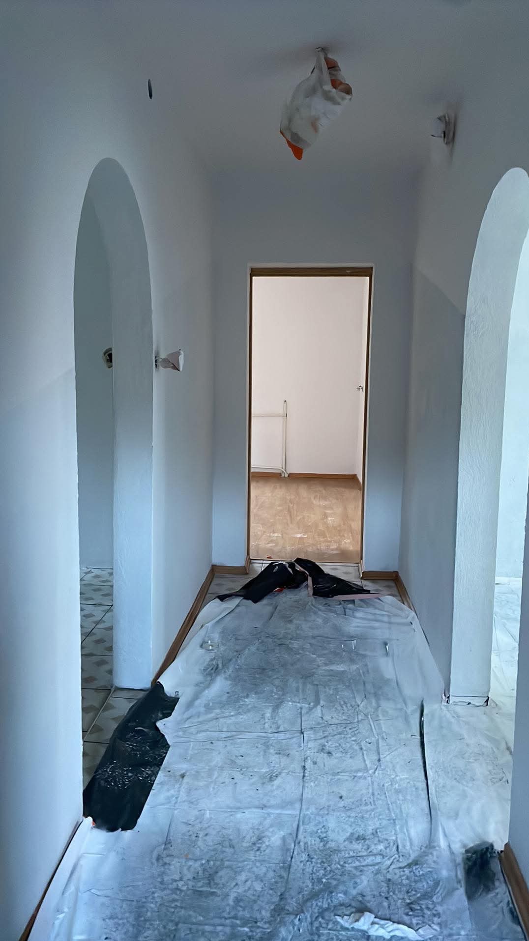 Korytarz w trakcie malowania: świeżo pomalowane ściany na biało, podłoga zabezpieczona folią malarską, widoczne łuki w przejściach i otwarte drzwi do pokoju.