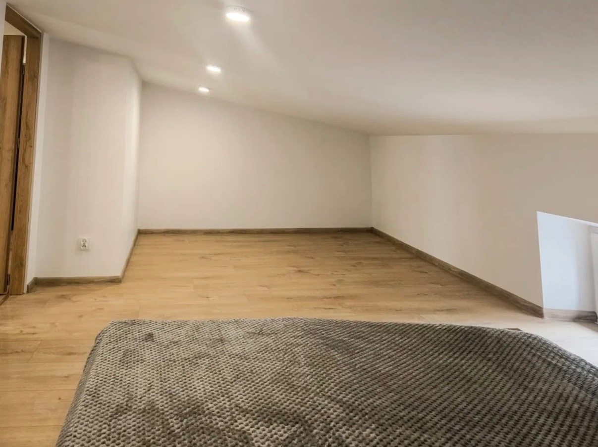 Poddasze z drewnianą podłogą i białymi ścianami, oświetlone trzema punktowymi lampami sufitowymi. W rogu widoczne drewniane drzwi, a na pierwszym planie fragment szarej narzuty.
