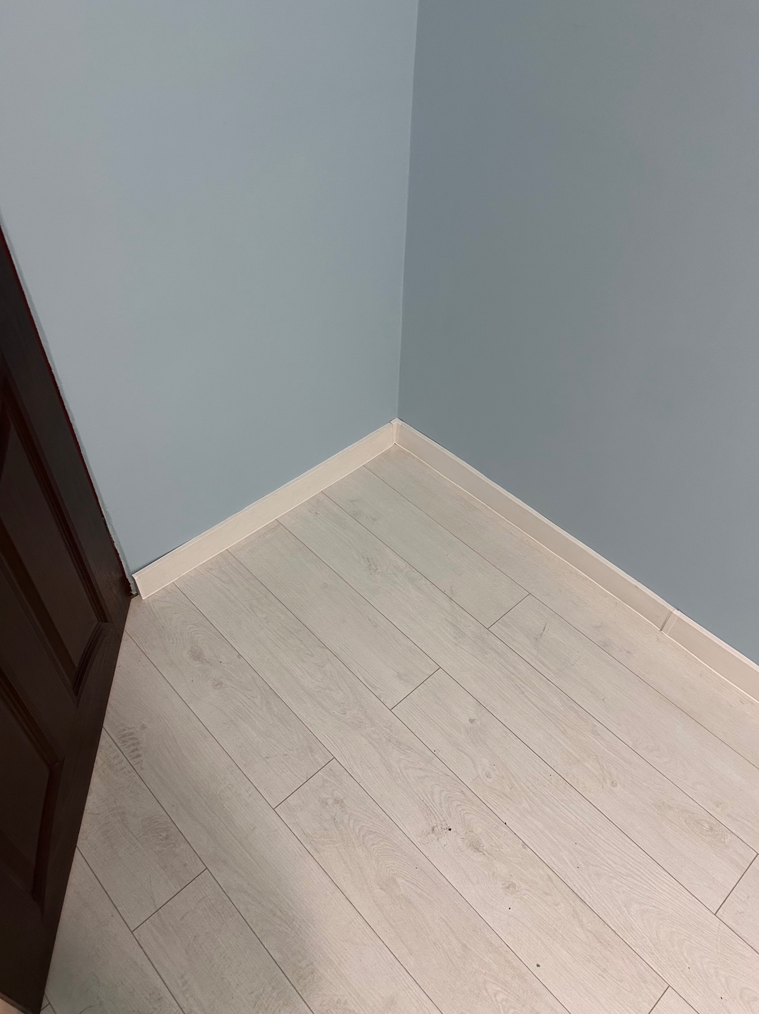 Jasne panele podłogowe ułożone w rogu pokoju z błękitnymi ścianami i brązowymi drzwiami. Widoczne białe listwy przypodłogowe.