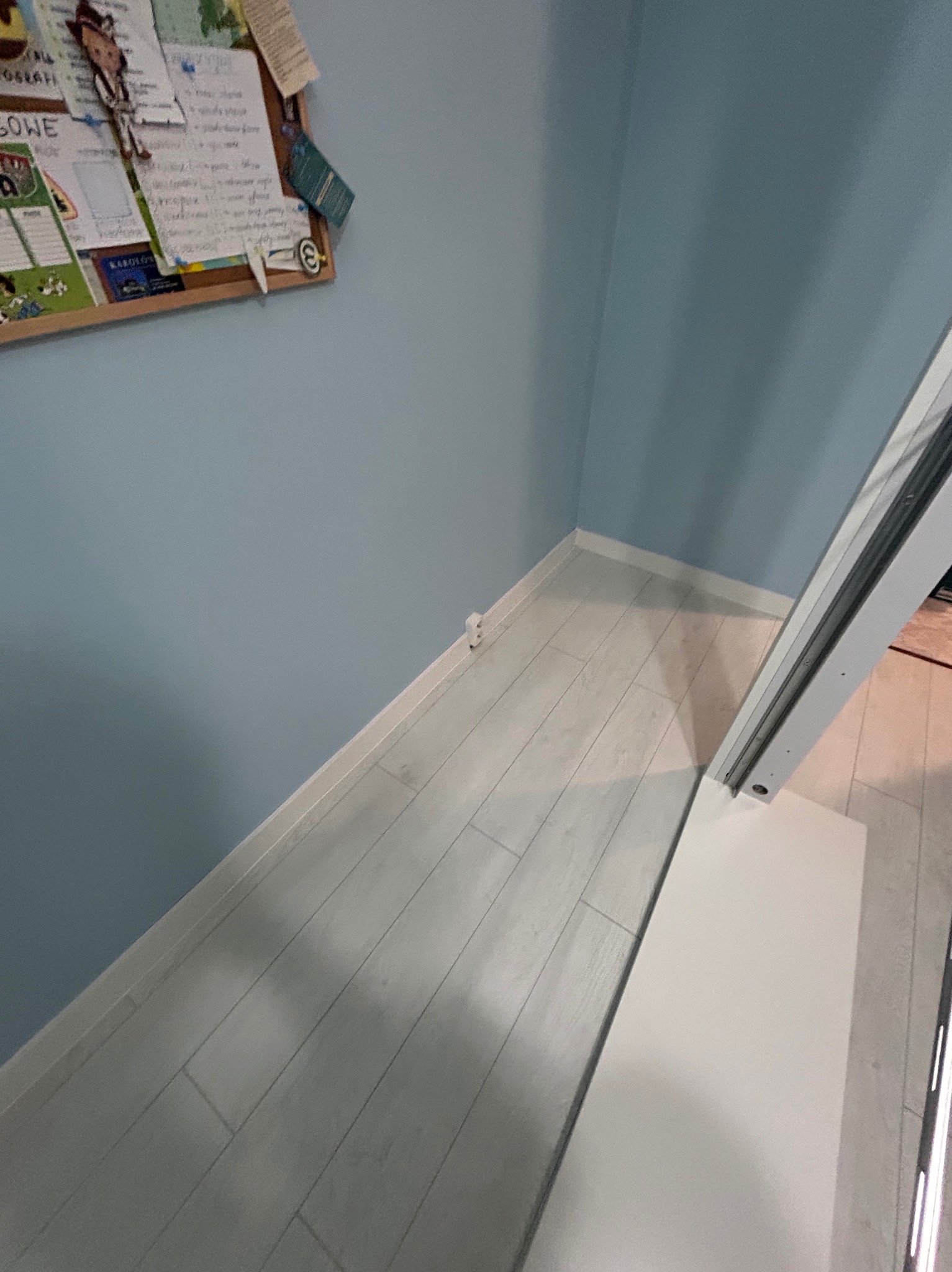 Jasne panele podłogowe ułożone wzdłuż ściany pomalowanej na błękitno. Widoczna listwa przypodłogowa i fragment białej konstrukcji meblowej.