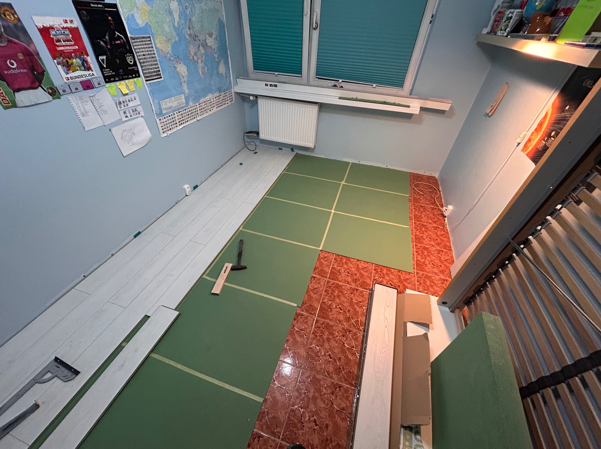 Fragment pokoju w trakcie układania paneli podłogowych. Widoczne narzędzia, podkład i częściowo ułożone panele w jasnym odcieniu drewna.