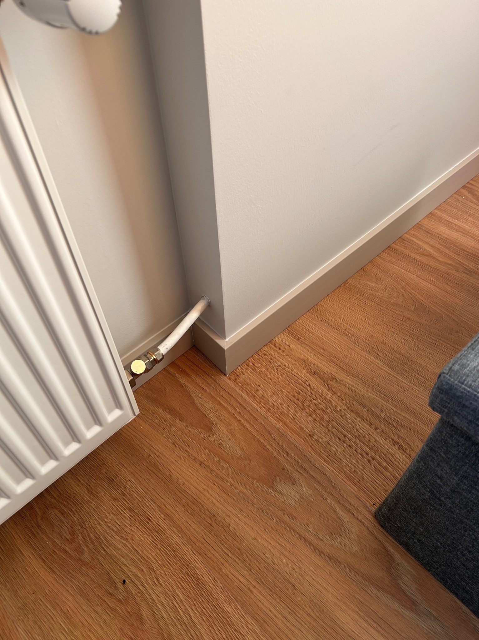 Biały grzejnik panelowy przy ścianie z listwą przypodłogową i drewnopodobną podłogą. Widoczne rury doprowadzające wodę do grzejnika.