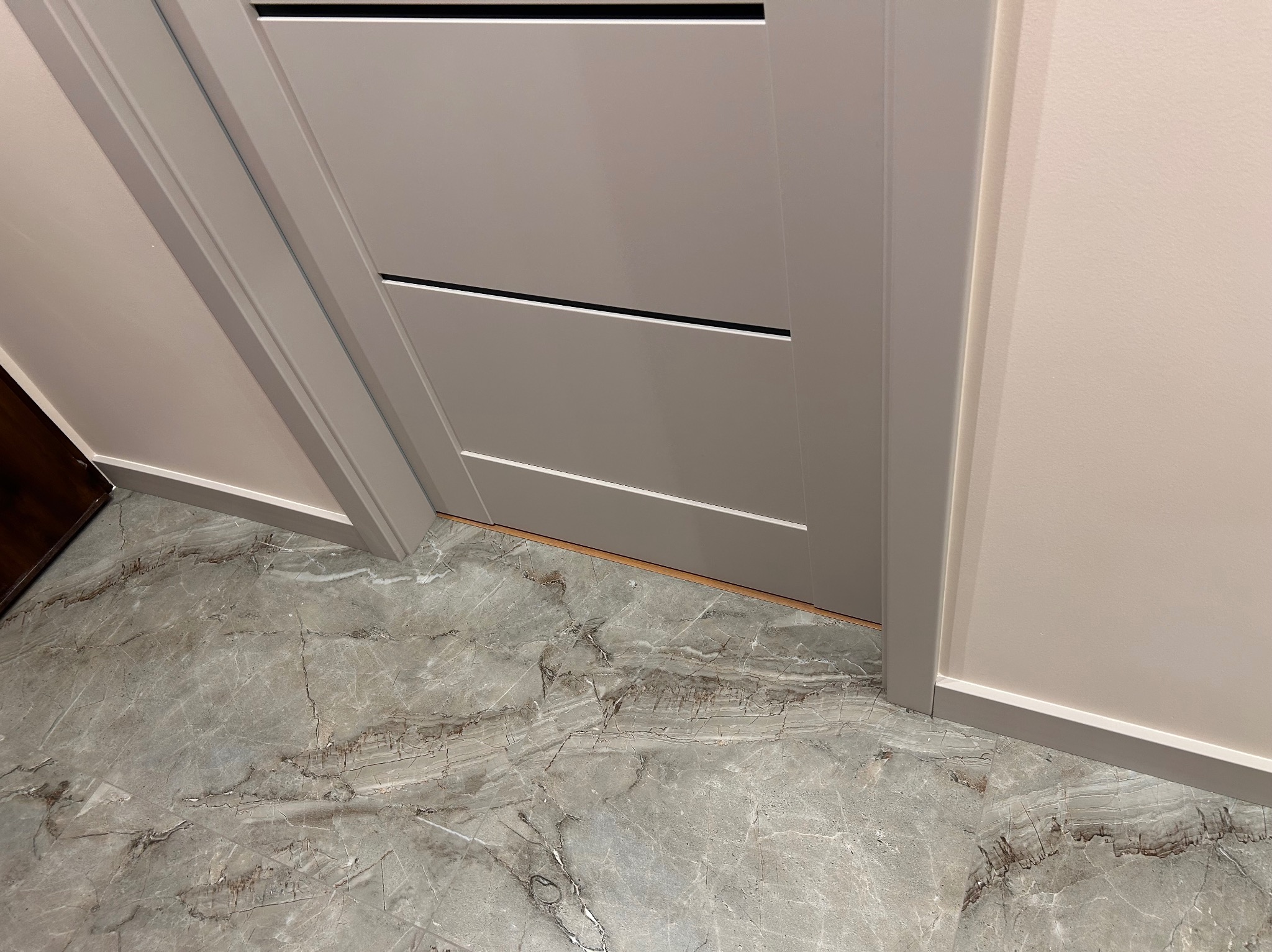 Zamknięte, szare drzwi wewnętrzne z czarnym akcentem, widoczne od dołu, z fragmentem marmurowej podłogi i kremowej ściany. Minimalistyczny design.