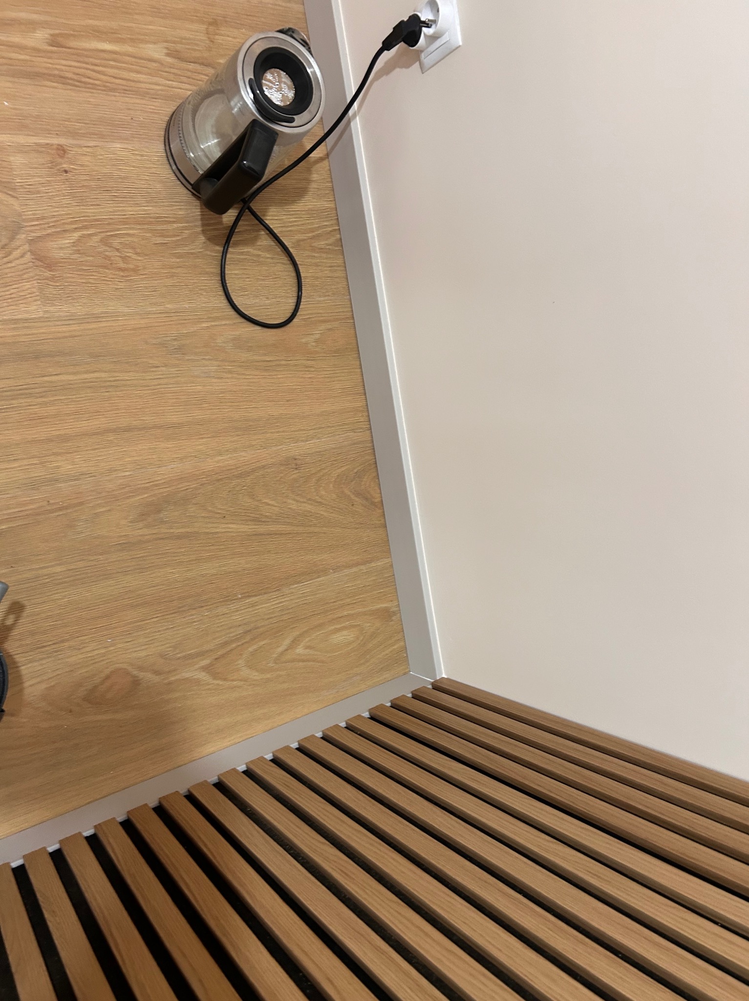 Nowoczesne wykończenie podłogi z drewnianych listew w jasnym odcieniu, kontrastujące z gładką, beżową ścianą i fragmentem paneli. Widoczny czajnik elektryczny.