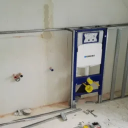 Stelaż podtynkowy Geberit zamontowany przy ścianie w trakcie prac wykończeniowych łazienki na poddaszu, widoczne instalacje i poziomica.