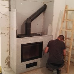 TymiRękami - Mężczyzna montuje obudowę kominka z płyt gipsowo-kartonowych. Widoczny czarny wkład kominkowy i rura dymowa. Na podłodze narzędzia i drabina.