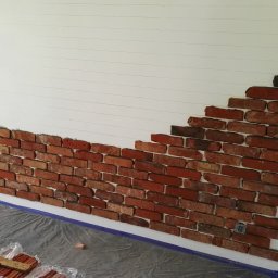 TymiRękami - Fragment ściany z dekoracyjną cegłą w trakcie montażu, widoczne linie pomocnicze i folia ochronna. Częściowo wykończona ściana w pomieszczeniu.