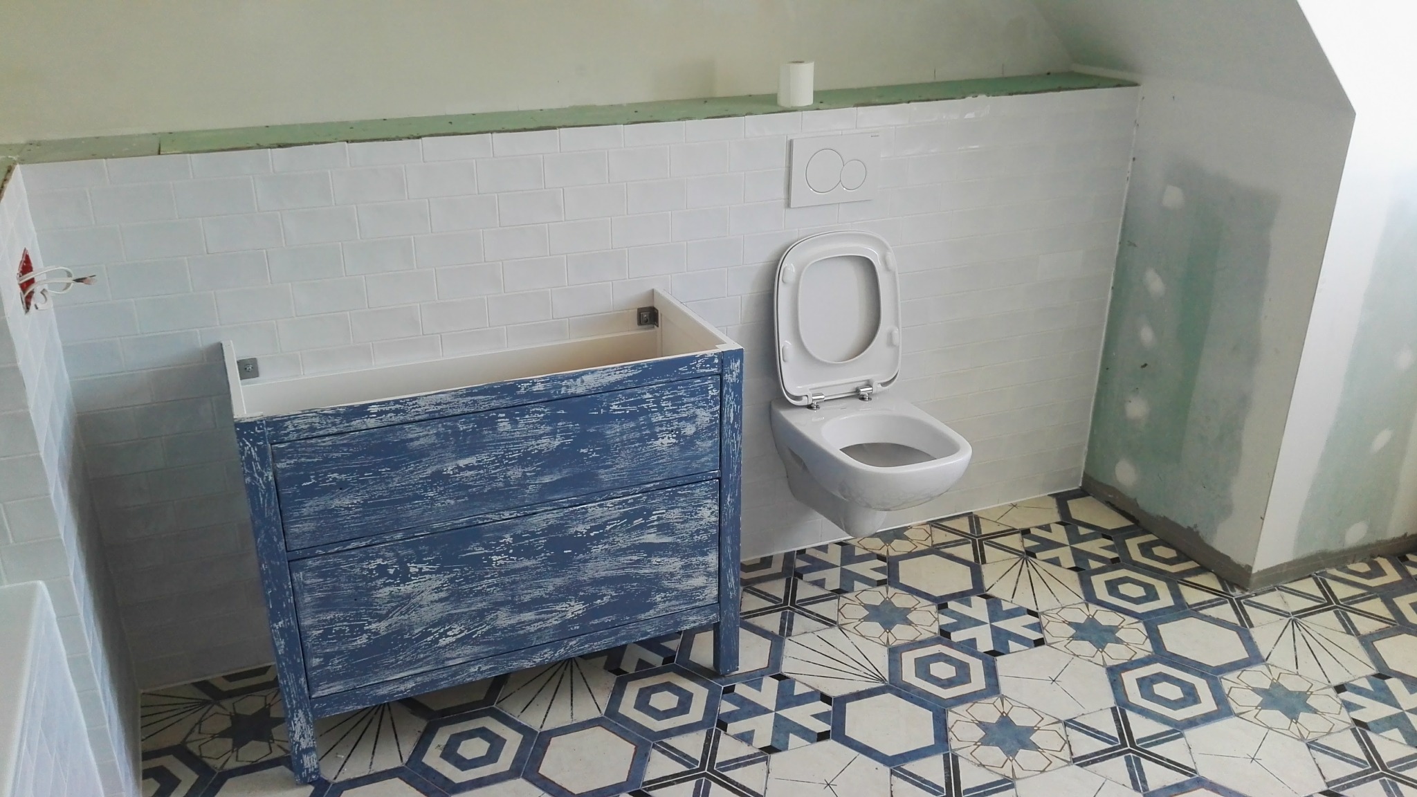 Łazienka w trakcie wykończenia: białe płytki na ścianach, wzorzysta podłoga, niebieska szafka pod umywalkę w stylu vintage i podwieszana toaleta.