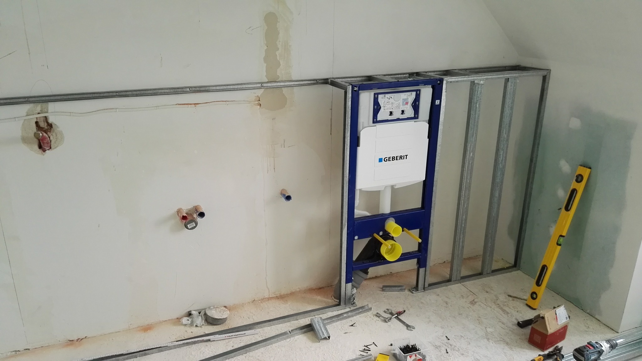 Stelaż podtynkowy Geberit zamontowany przy ścianie w trakcie prac wykończeniowych łazienki na poddaszu, widoczne instalacje i poziomica.