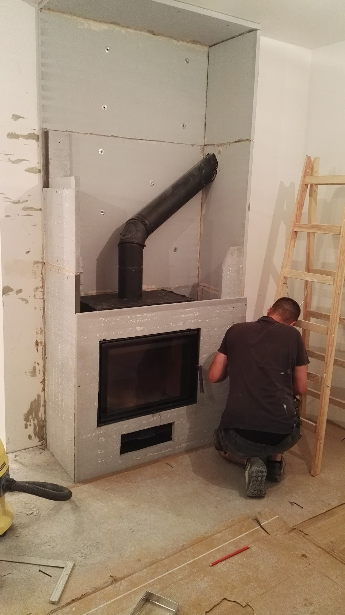 Mężczyzna montuje obudowę kominka z płyt gipsowo-kartonowych. Widoczny czarny wkład kominkowy i rura dymowa. Na podłodze narzędzia i drabina.