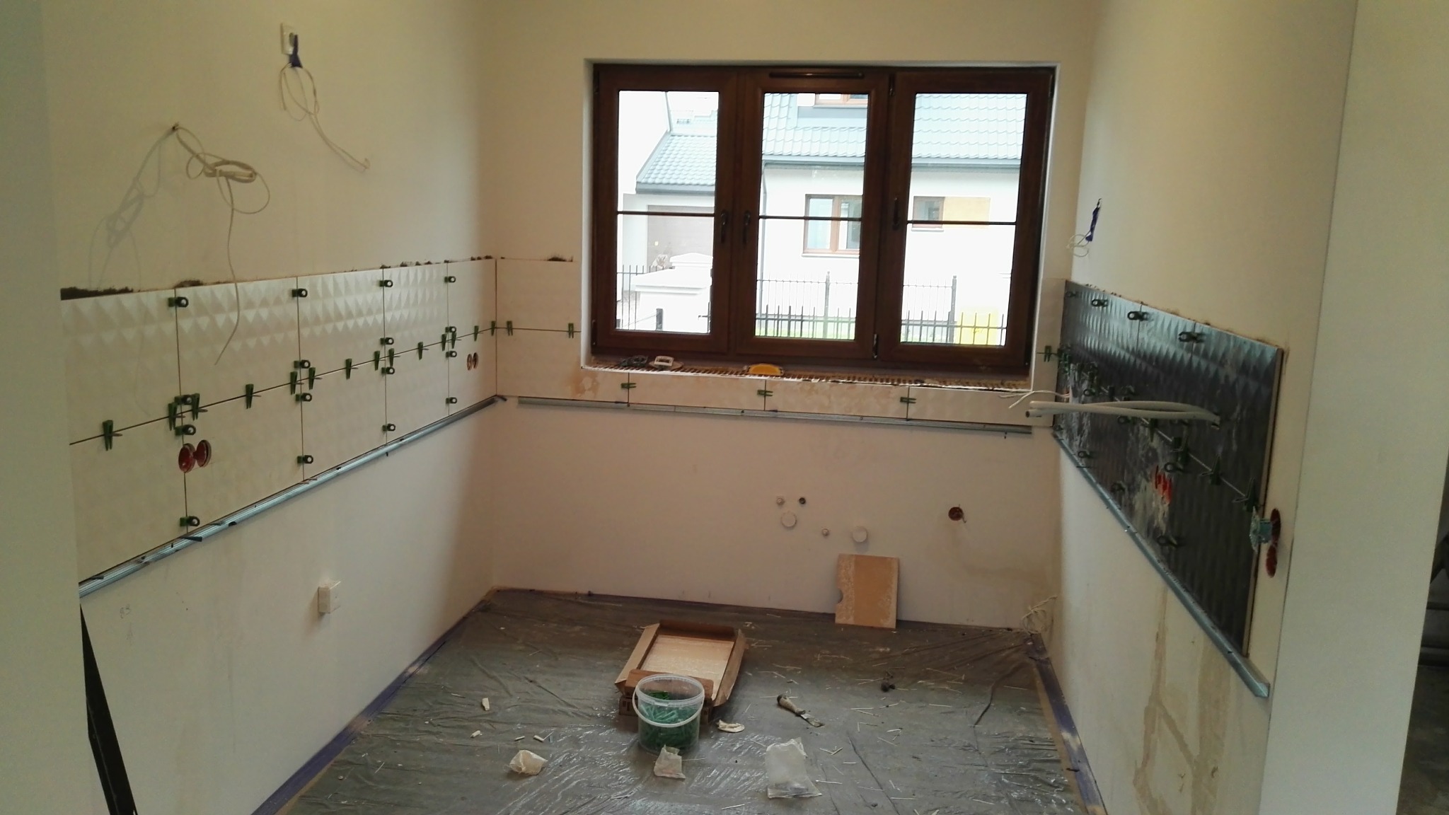 Wnętrze w trakcie układania płytek z poziomowaniem. Widoczne metalowe profile, krzyżyki dystansowe i przewody elektryczne. Okno z drewnianą ramą. Folia ochronna na podłodze.
