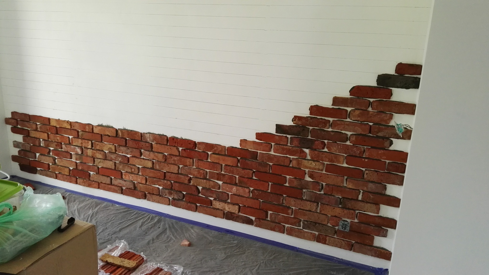 Fragment ściany z dekoracyjną cegłą w trakcie montażu, widoczne linie pomocnicze i folia ochronna. Częściowo wykończona ściana w pomieszczeniu.
