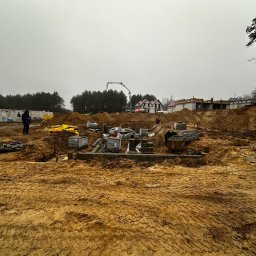 OkhremBud - Fundamenty domu w trakcie budowy, widoczne bloczki fundamentowe, ziemia i sprzęt budowlany. W tle las i częściowo wybudowane ściany.