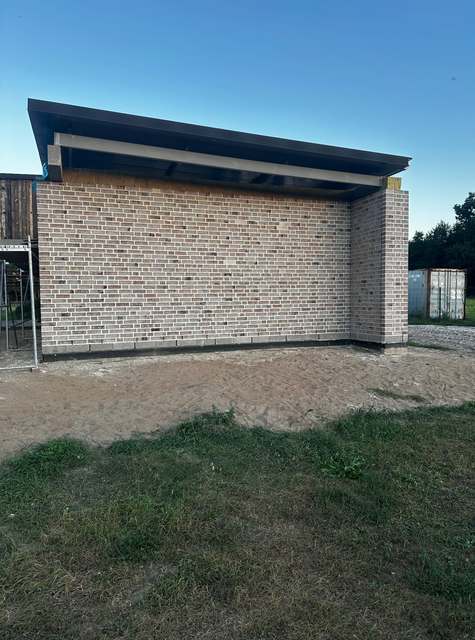 Ściana budynku z szarej cegły, zadaszenie z elementami stalowymi. Widoczny fragment rusztowania i kontener. Teren wokół budynku z piaskiem i trawą.